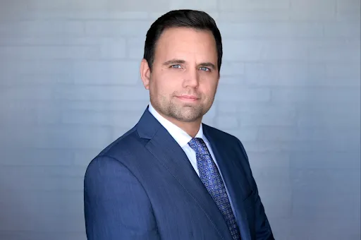 Professional portrait of a man in a blue suit and patterned tie against a gray background.