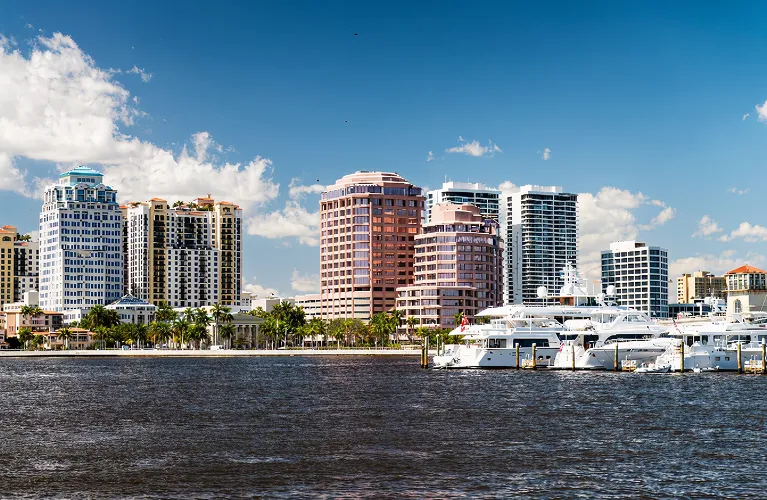 Waterfront cityscape with modern high-rise buildings, palm trees, and luxury yachts under a blue sky.