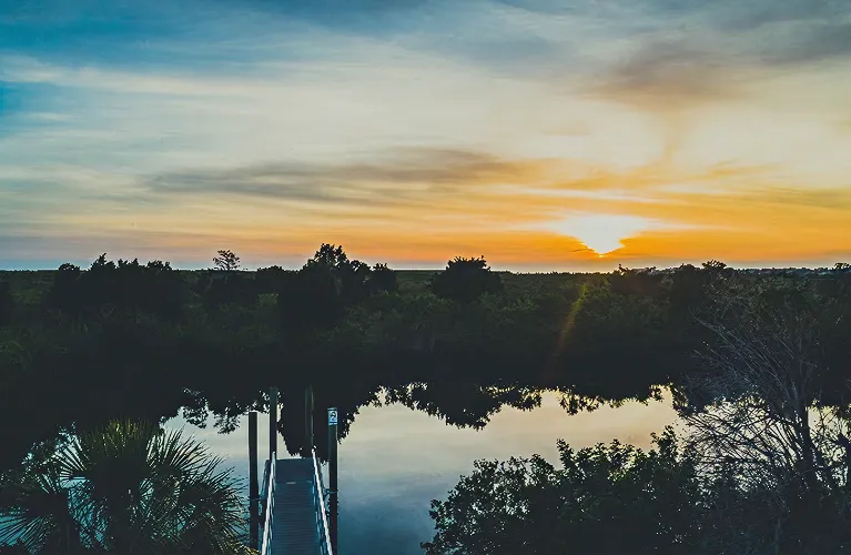 Sunset over calm water with silhouetted trees and a dock extending into the water.