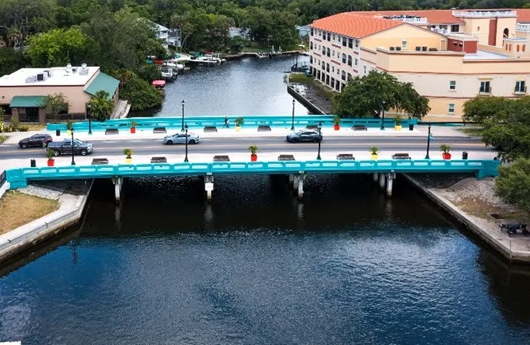Aerial view of a turquoise bridge over a river with cars, benches, planters, and surrounding buildings.