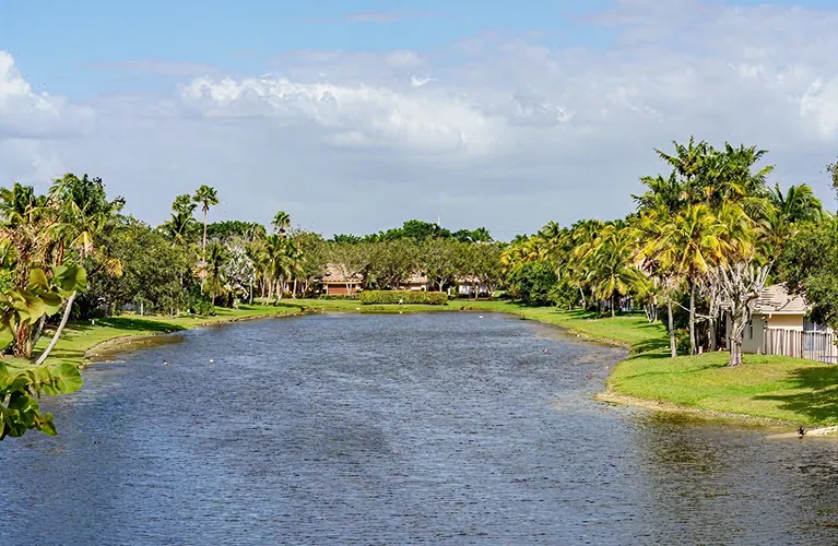 Calm river flanked by green palm trees and residential houses under a partly cloudy sky.
