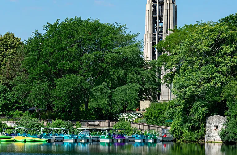 Calm lake with colorful paddle boats docked, surrounded by lush green trees and a tall stone tower.
