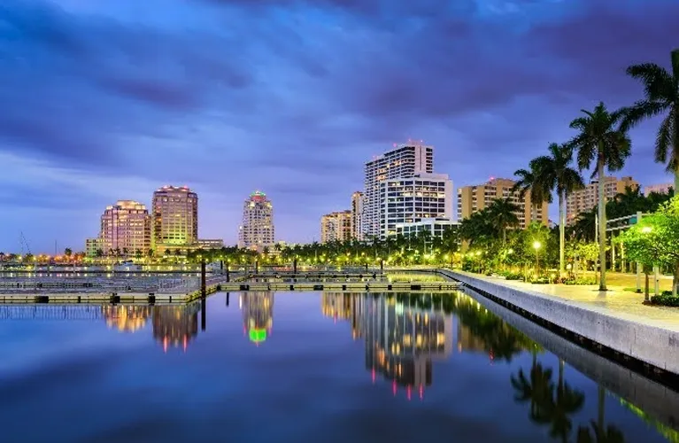 Evening cityscape with illuminated buildings, palm trees, and waterfront reflecting lights.
