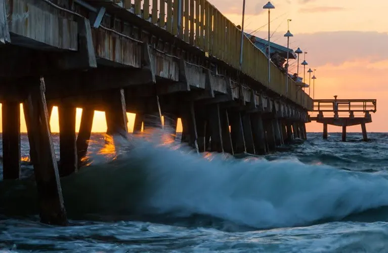 Wooden pier with lampposts over turbulent ocean waves at sunset.