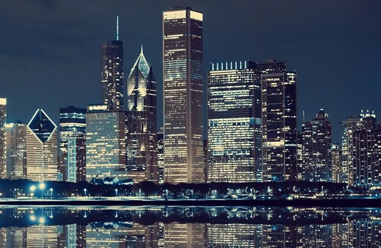 Nighttime city skyline with illuminated skyscrapers reflected on calm water.