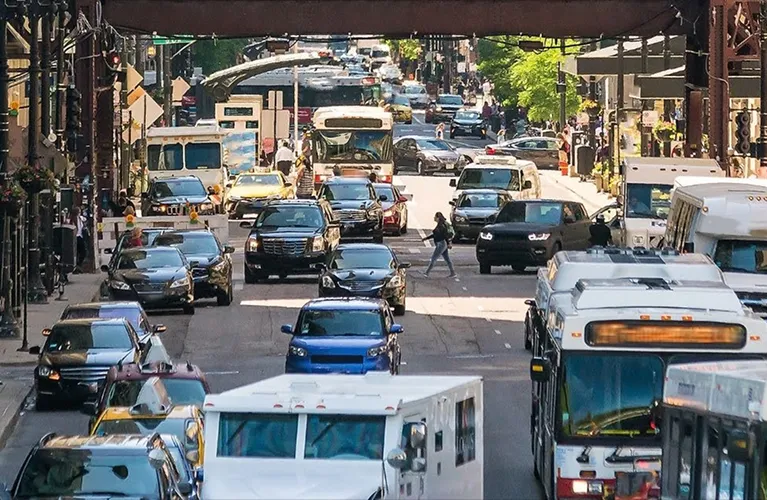 Busy urban street with multiple vehicles, including cars, buses, and pedestrians crossing the road.