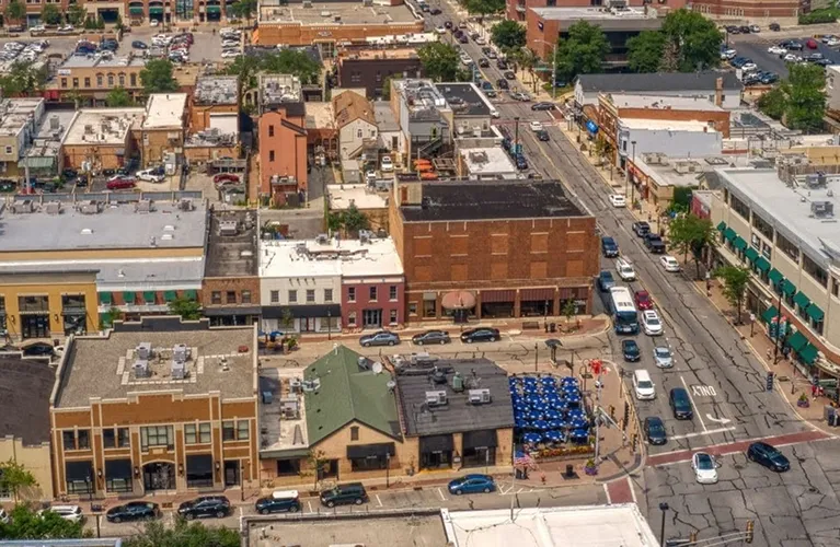 Aerial view of a busy urban intersection with commercial buildings, parked cars, and outdoor seating with blue umbrellas.