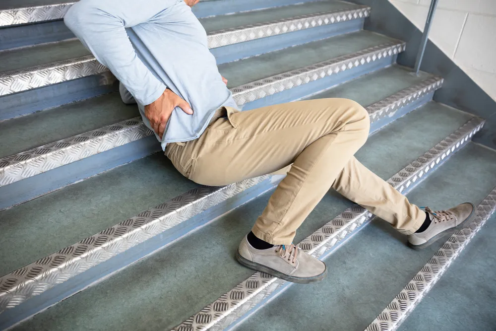 Adult man in beige pants clutching lower back, sitting on metal stairs in pain.