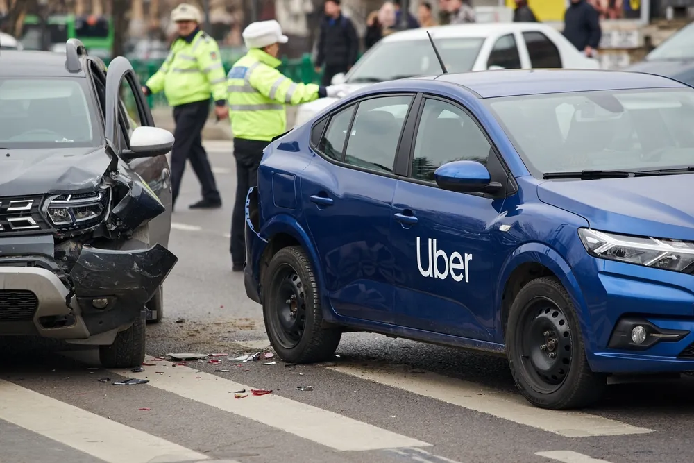 Two damaged cars, including a blue Uber vehicle, on a street with police officers in high-visibility jackets in the background.