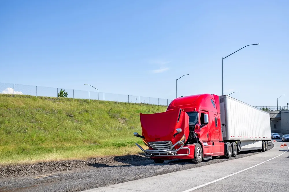 Red semi-truck with raised hood parked on highway shoulder, warning triangles placed behind.
