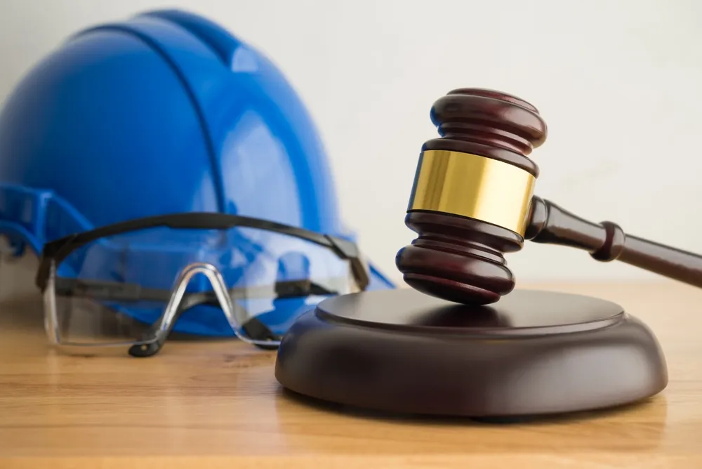 Wooden judge's gavel with gold band, blue construction helmet, and safety glasses on wooden surface.