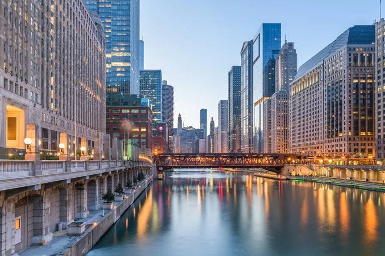 Evening cityscape of a river flanked by illuminated high-rise buildings and a bridge.