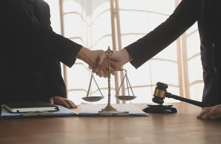 Two professionals shaking hands over a desk with legal scales and a gavel.